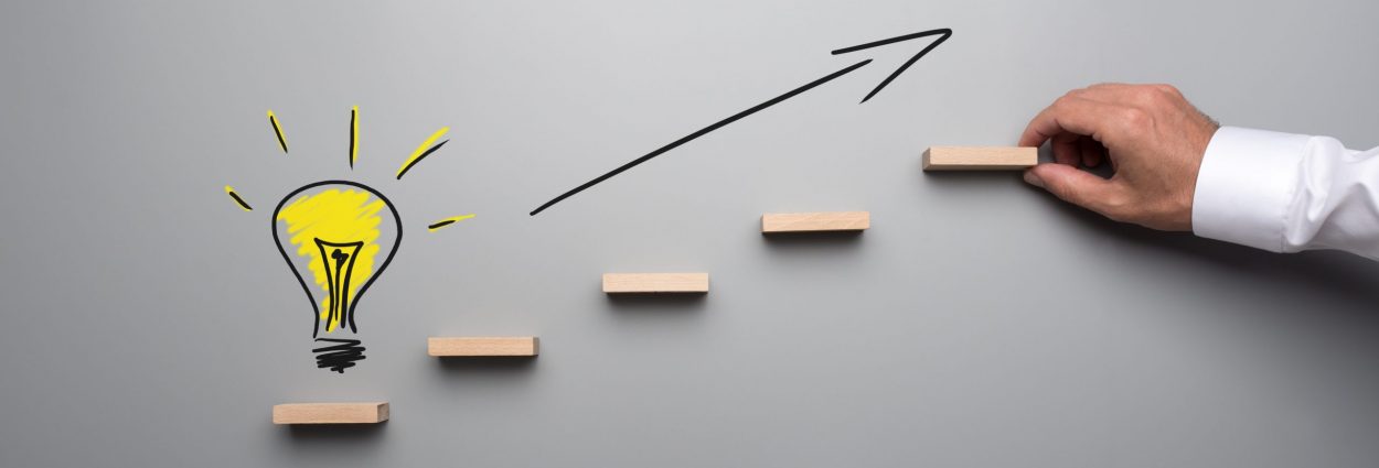 Yellow hand drawn light bulb on the bottom of wooden steps with black arrow pointing upwards in a conceptual image of idea and success.